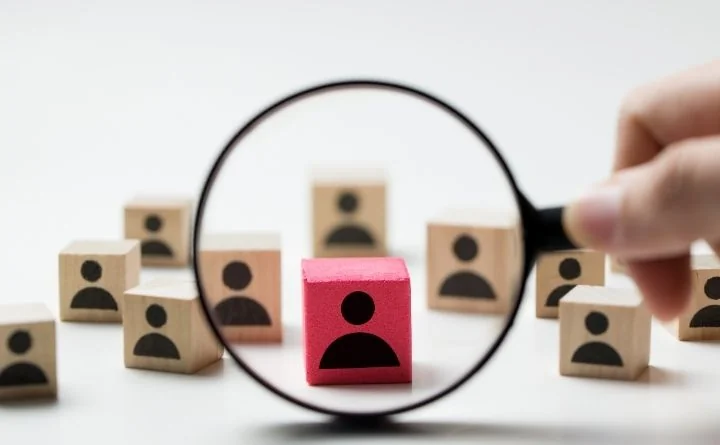 Lupa skupiona na czerwonym klocku z ikoną osoby wśród drewnianych klocków z sylwetkami.