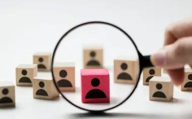 Lupa skupiona na czerwonym klocku z ikoną osoby wśród drewnianych klocków z sylwetkami.