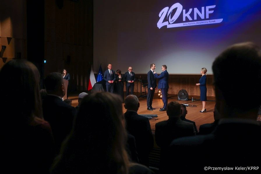 Uroczystość z okazji 20-lecia KNF na scenie audytorium, podczas której mężczyzna wręcza odznaczenie innemu mężczyźnie w obecności oficjeli i publiczności.