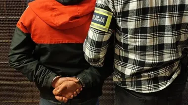 Funkcjonariusz policji w kraciastej koszuli prowadzi zatrzymanego w kajdankach mężczyznę w czarno-czerwonej kurtce.