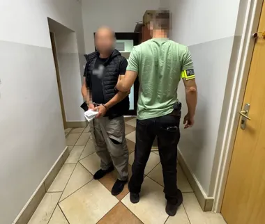 Funkcjonariusz policji w zielonej koszulce prowadzi zatrzymanego mężczyznę w kajdankach przez korytarz budynku.