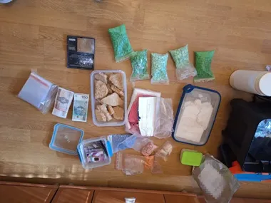 Na drewnianym stole ułożono zabezpieczone przez służby substancje, tabletki, pieniądze, wagę i opakowania związane z przestępczością narkotykową.
