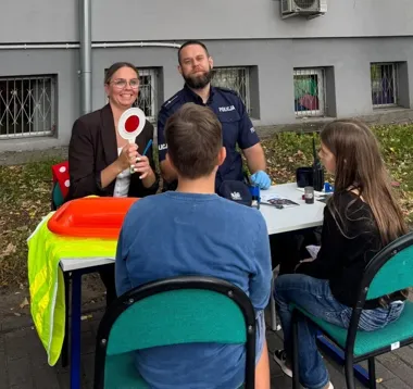 Policjant i kobieta rozmawiają z dziećmi przy stole podczas wydarzenia edukacyjnego na zewnątrz.