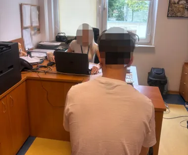 Mężczyzna siedzi przy biurku urzędniczki w pomieszczeniu biurowym, kobieta pracuje przy laptopie.