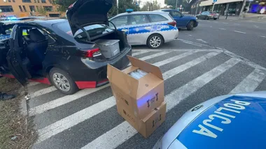 Policyjna akcja na przejściu dla pieszych: radiowozy otaczają czarny sedan z otwartym bagażnikiem, przy którym stoją kartony.