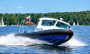 Policyjna łódź motorowa z napisem POLICJA płynie po jeziorze w słoneczny dzień, w tle widać lasy i żaglówki.