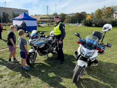 Policjant w kamizelce odblaskowej rozmawia z dwójką dzieci stojących przy policyjnych motocyklach na trawiastym terenie podczas plenerowej imprezy.