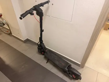 Czarna elektryczna hulajnoga stoi w korytarzu na kafelkowej podłodze przy ścianie z szafkami technicznymi.