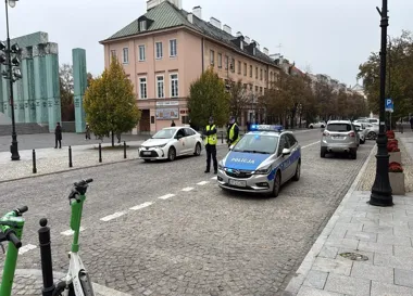 Policjanci w kamizelkach odblaskowych stoją przy policyjnym radiowozie na brukowanej ulicy w historycznej części miasta.