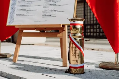 Przed drewnianą sztalugą z dokumentem stoi mosiężny wazon z biało-czerwoną wstążką, a obok widoczne są czerwone flagi na brukowanej nawierzchni.