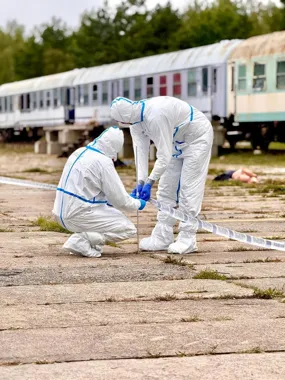 Dwie osoby w kombinezonach ochronnych zabezpieczają teren taśmą policyjną na opuszczonej stacji kolejowej.
