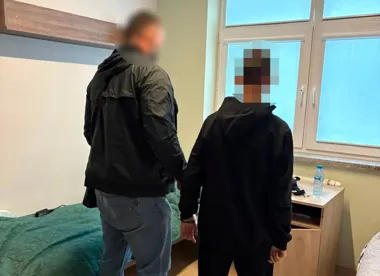 Dwie osoby w ciemnych kurtkach stoją w pokoju z łóżkiem i oknem, jedna z nich ma zasłoniętą twarz.