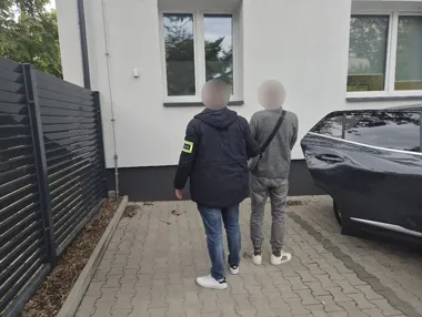 Funkcjonariusz w kurtce z żółtą opaską na rękawie prowadzi mężczyznę w szarej bluzie w stronę budynku przy zaparkowanym czarnym samochodzie.