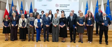 Grupa osób z dyplomami pozuje do zdjęcia podczas uroczystości Dnia Edukacji Narodowej z napisem „Tytuł Honorowy Przodownika Oświaty” na tle.