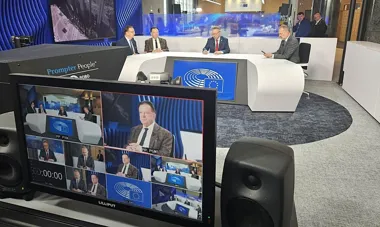 Czterech mężczyzn w garniturach siedzi przy białym, owalnym stole w studiu telewizyjnym z logo Parlamentu Europejskiego, a w pierwszym planie widać monitor reży
