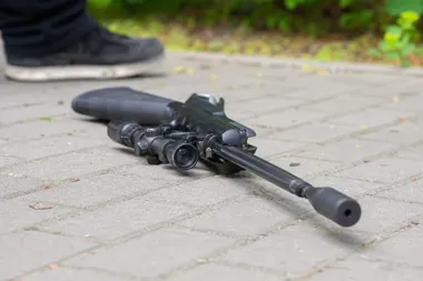 Karabin z celownikiem optycznym leży na brukowanej nawierzchni, w tle widoczna stopa osoby w czarnym obuwiu.