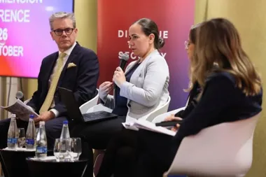 Trzy osoby uczestniczą w panelu dyskusyjnym podczas konferencji, jedna z nich przemawia do mikrofonu.