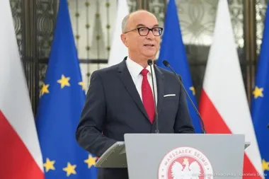 Mężczyzna w garniturze przemawia z pulpitu z herbem Marszałka Sejmu, w tle flagi Polski i Unii Europejskiej.