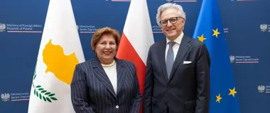 Kobieta i mężczyzna w strojach biznesowych pozują do wspólnego zdjęcia na tle flag Cypru, Polski i Unii Europejskiej.