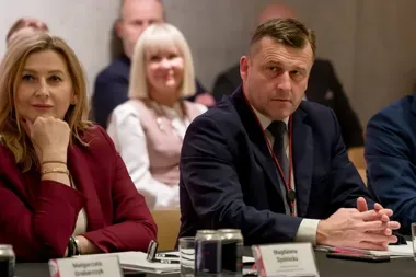 Uczestnicy konferencji lub spotkania biznesowego siedzą przy stole z tabliczkami z nazwiskami, puszkami napojów i dokumentami.
