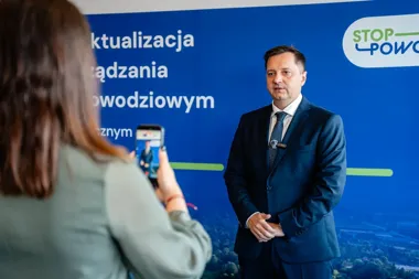 Mężczyzna w garniturze stoi przed niebieską ścianką z napisami dotyczącymi powodzi, a kobieta nagrywa go smartfonem.