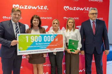 Grupa osób na tle czerwonej ścianki z logo Mazowsze trzyma symboliczny czek na 1 900 000 zł dla Miasta Łaskarzew na przebudowę bieżni lekkoatletycznej oraz bois