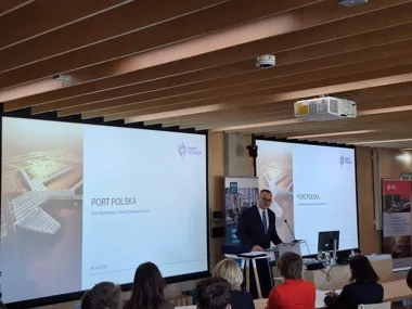 Mężczyzna w garniturze przemawia zza pulpitu podczas konferencji w sali z drewnianym sufitem, za nim dwa ekrany z prezentacją „PORT POLSKA”.