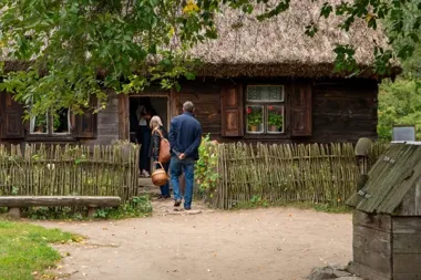 Trzy osoby wchodzą do drewnianej chaty z dachem z trzciny, otoczonej płotem z wikliny, w skansenie lub zabytkowym gospodarstwie wiejskim.