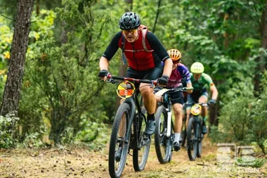 Trzech mężczyzn w kaskach i sportowych strojach jedzie na rowerach górskich leśną ścieżką podczas wyścigu lub zorganizowanej jazdy terenowej.