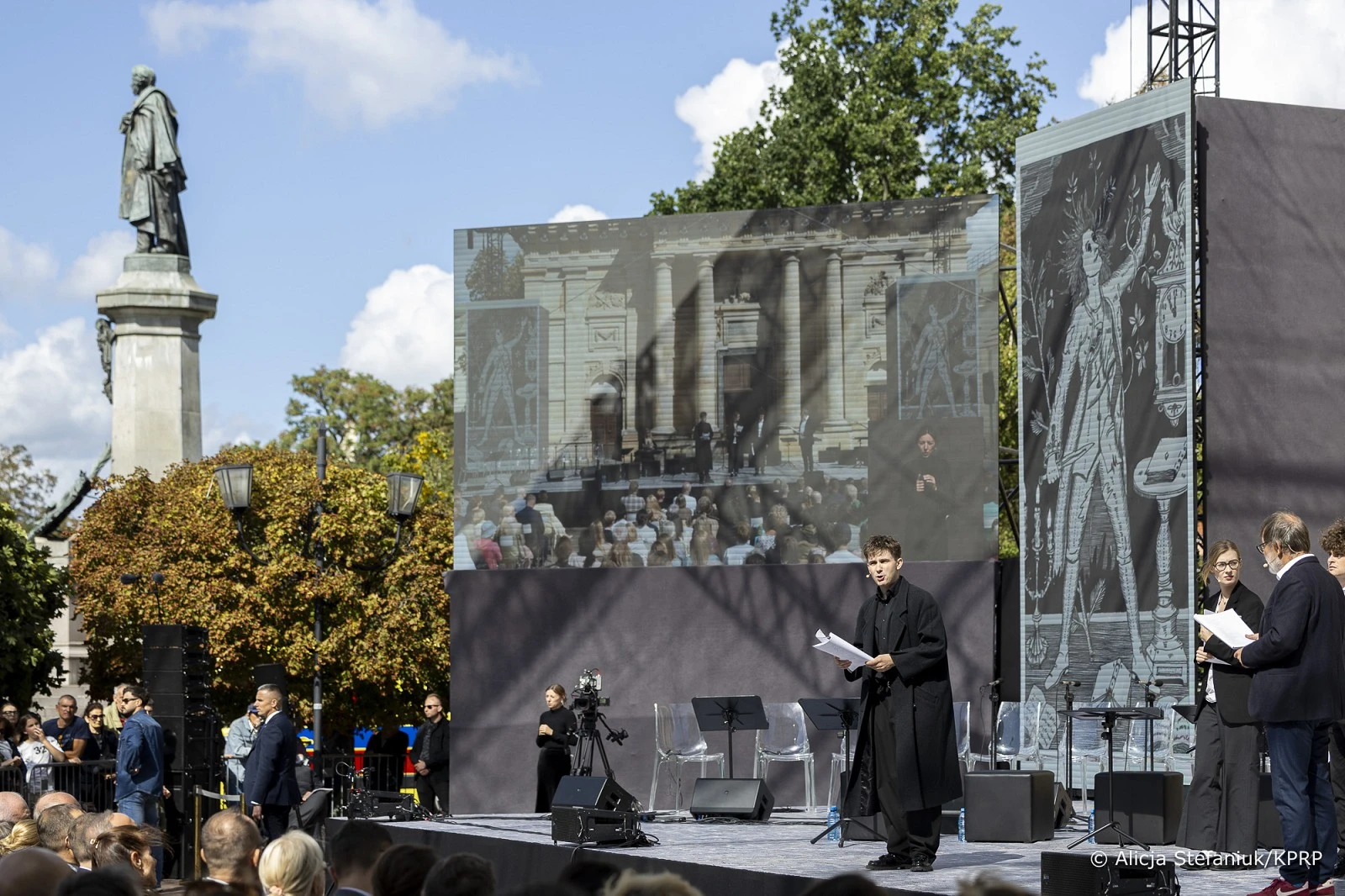 Mężczyzna w czarnym płaszczu czyta z kartki na scenie plenerowej przed ekranami i pomnikiem, w tle widownia i drzewa.